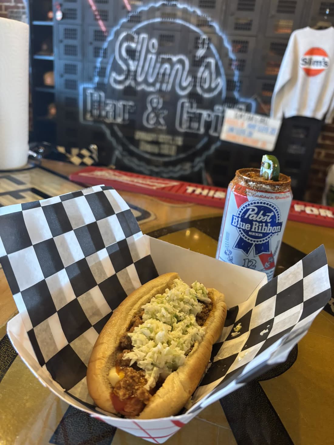 Chili slaw dog in a checkered paper basket with a dressed can of Pabst, the Slim's neon glowing behind the bar
